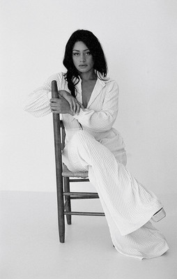 A woman wearing a white jumpsuit sits on a chair which is turned sideways, facing camera with her legs crossed and her arms relaxed on the chairback.