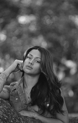A woman leans on a tree branch wearing an open jean jacket and showing cleavage as her hair cascades over her breasts.