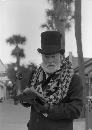 An older, bearded gentleman in a top hat and Victorian clothing stares down the camera as he holds an open book in his hands.