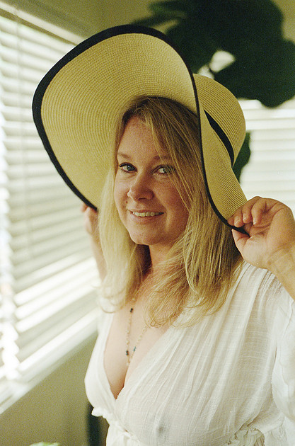 A beautiful woman looks sideways smiling at the camera, wearing a sheer white dress and a straw hat. Her deep neckline shows off her breasts and a hard nipple pokes through the material.