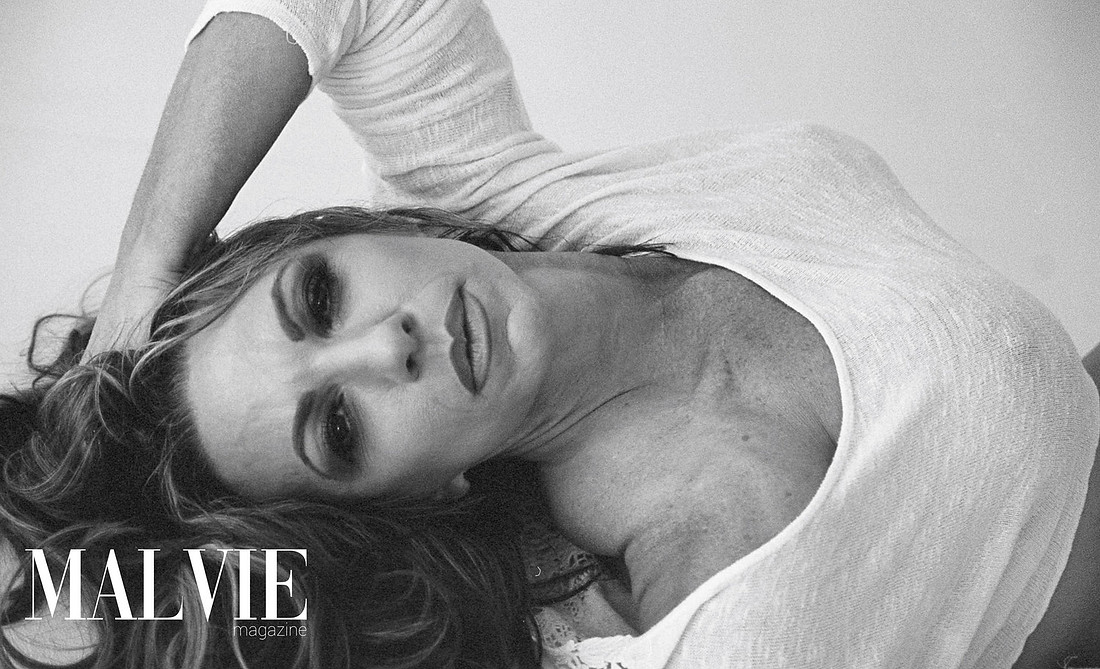 A beautiful woman lays sideways facing the camera. She is wearing a thin sweater with her nipple visible through it. Her left hand is up in her hair.